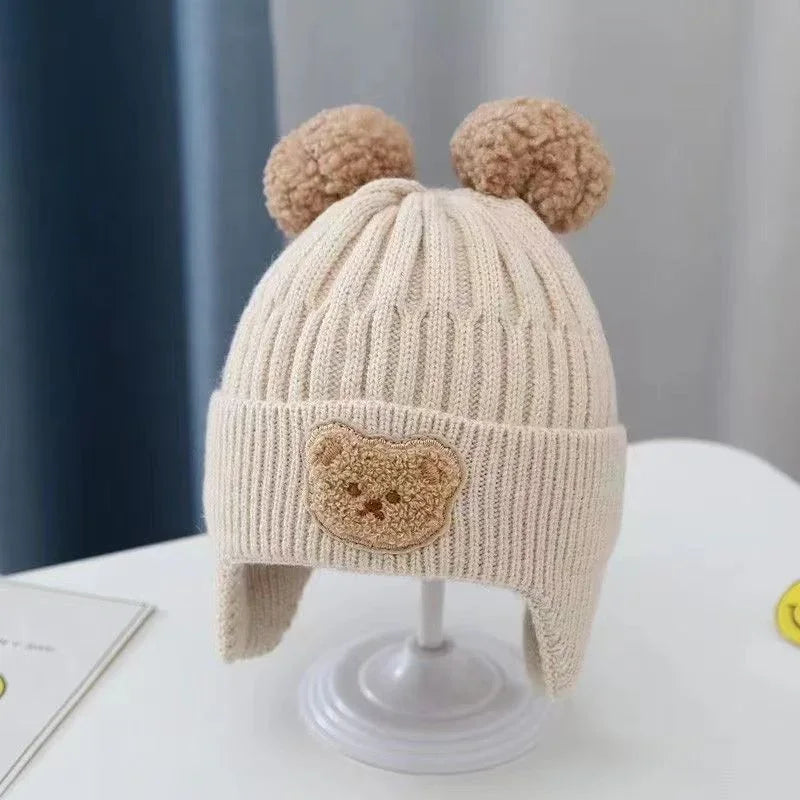 Beige knitted baby winter hat with bear ears pom-poms and teddy face patch on white stand