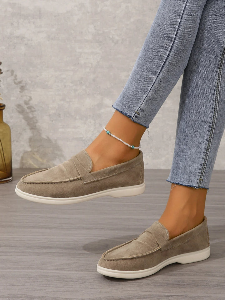 Woman wearing beige suede loafers and light blue jeans with a beaded ankle bracelet on wooden floor