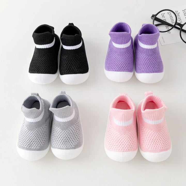 Four pairs of knitted baby sock shoes in black, purple, gray, and pink with white soles on white background