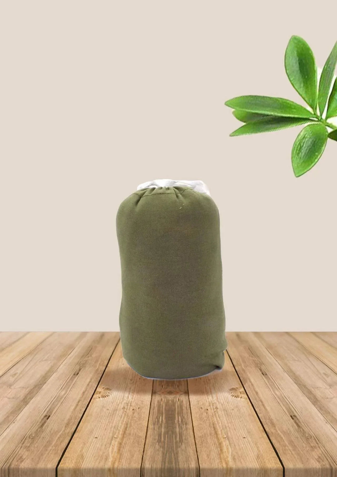 Compact olive green sleeping bag on a wooden floor with a green leaf branch on beige background