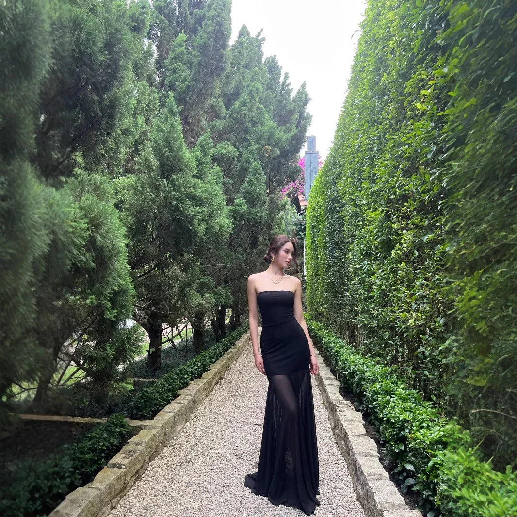 Woman in elegant black strapless evening gown standing on gravel path between dense green hedges and trees