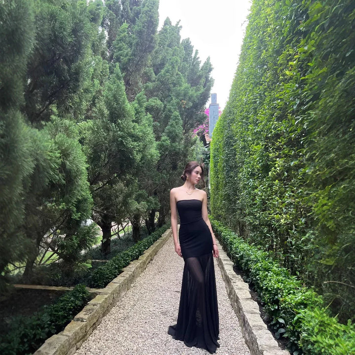 Woman in elegant black strapless evening gown standing on gravel path between dense green hedges and trees