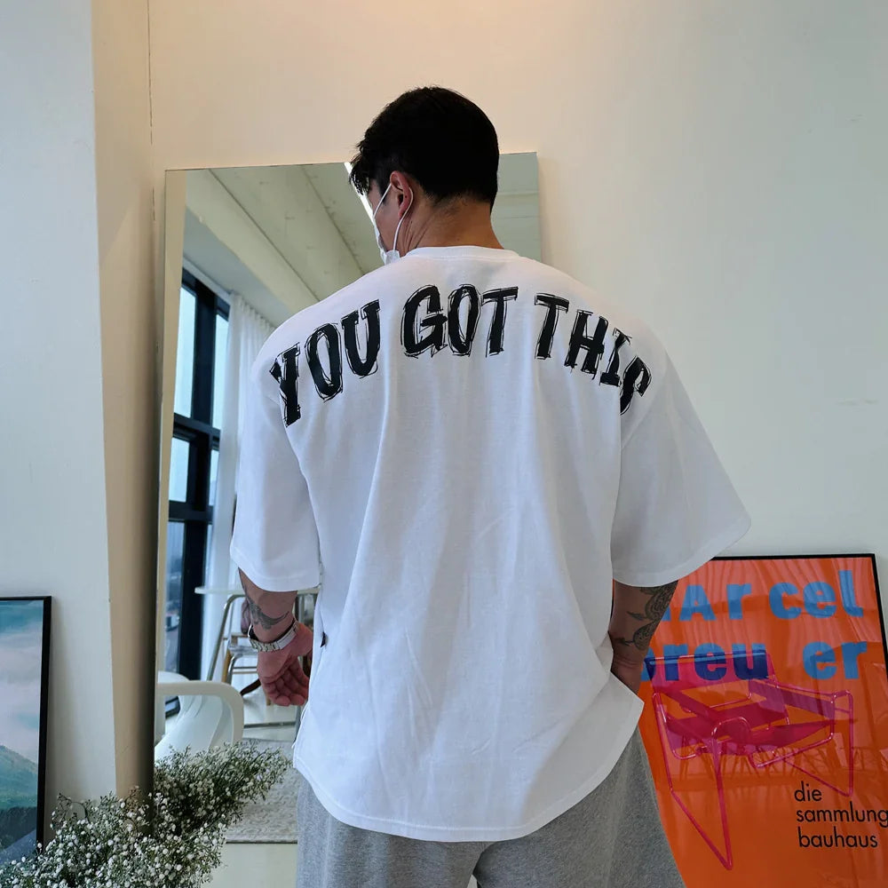 Man wearing white T-shirt with 'YOU GOT THIS' text, standing in a modern room with posters and mirror