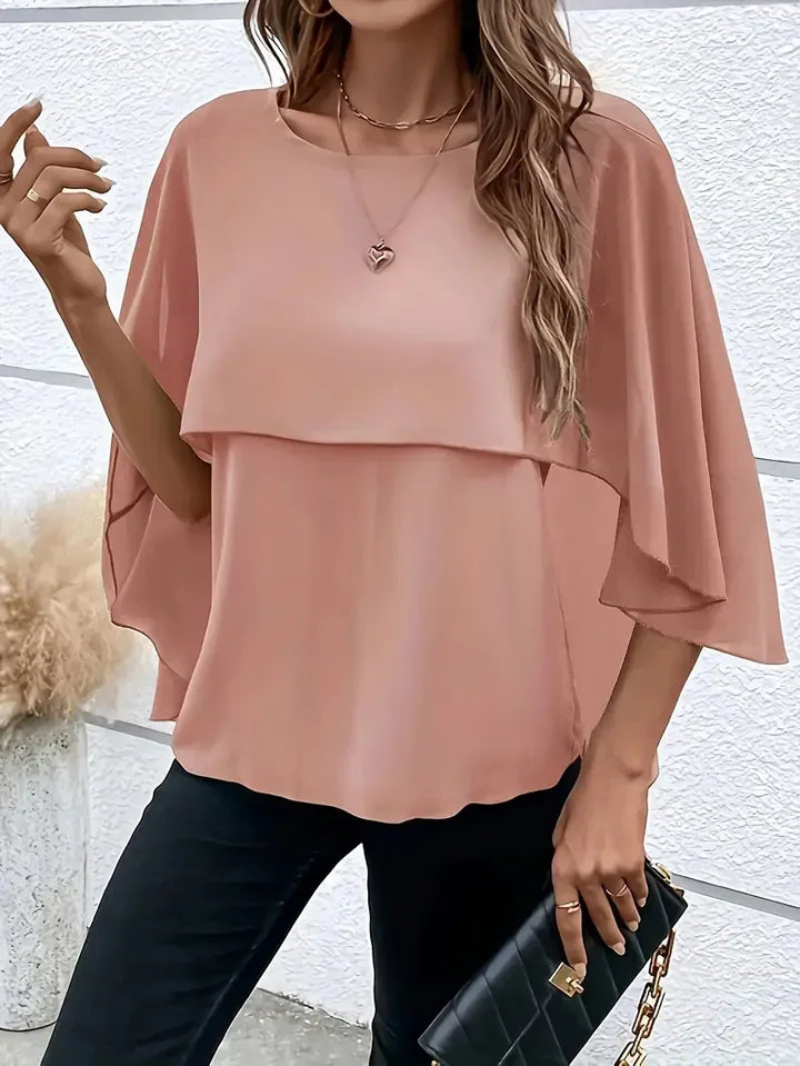 Woman wearing a peach chiffon layered blouse with flowy sleeves, black pants, and holding a black quilted clutch