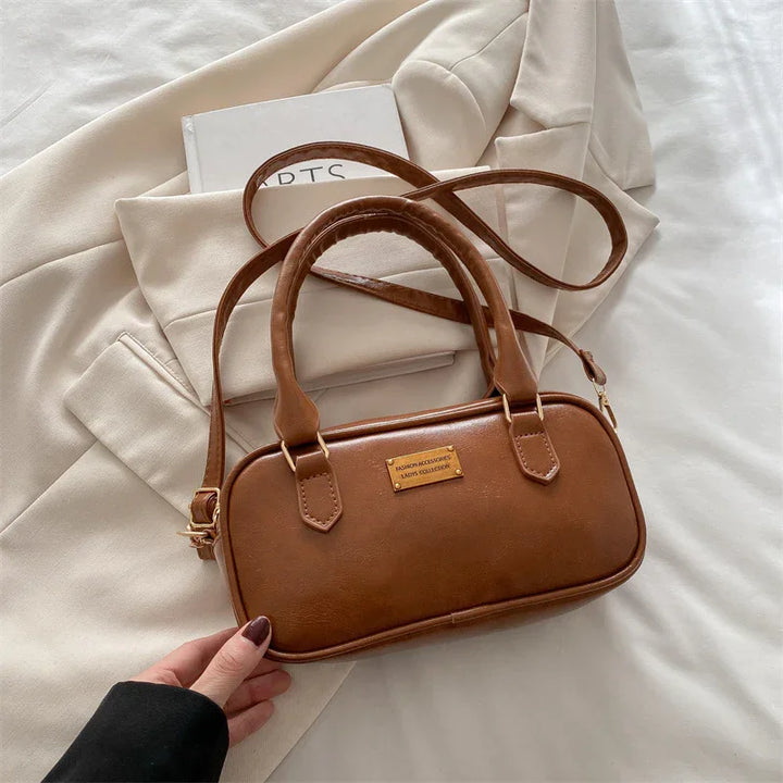 Brown leather handbag with dual handles and gold plaque on beige coat and white book background
