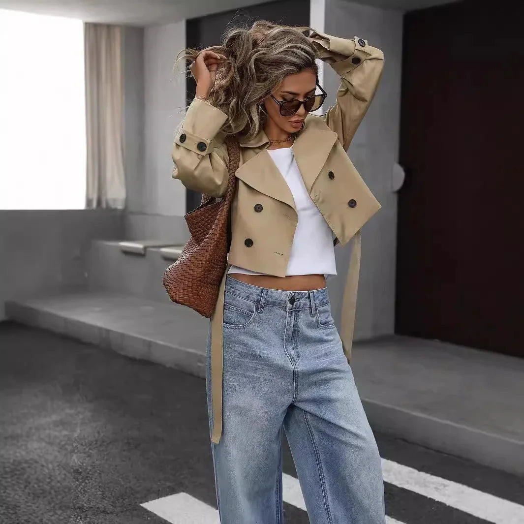Woman in beige cropped trench coat, white top, blue jeans, sunglasses, and brown woven bag outdoors