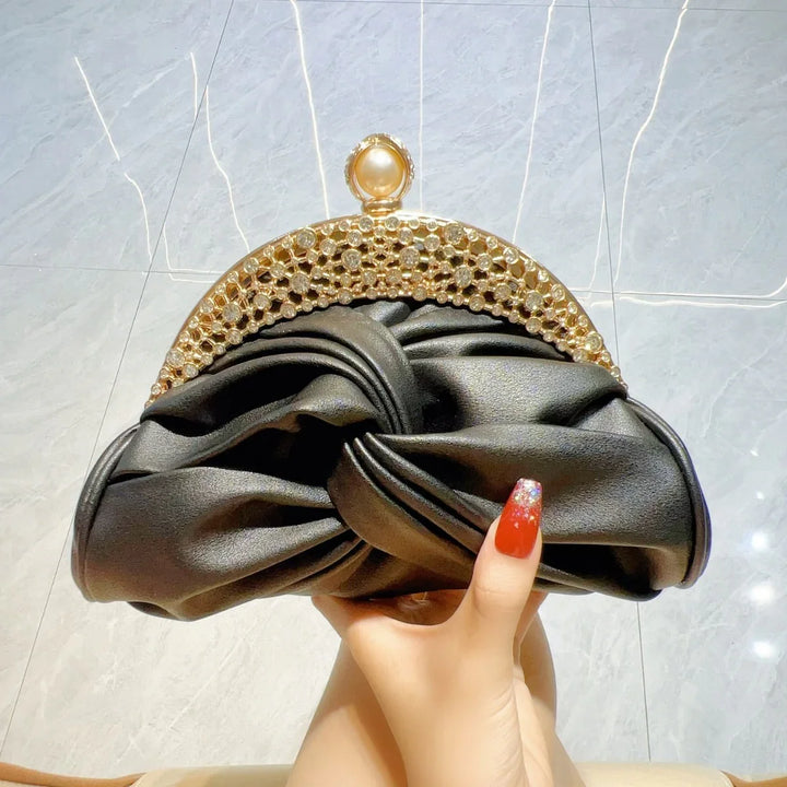 Black ruched satin clutch purse with gold jeweled frame and pearl clasp held by hand with red glitter nails