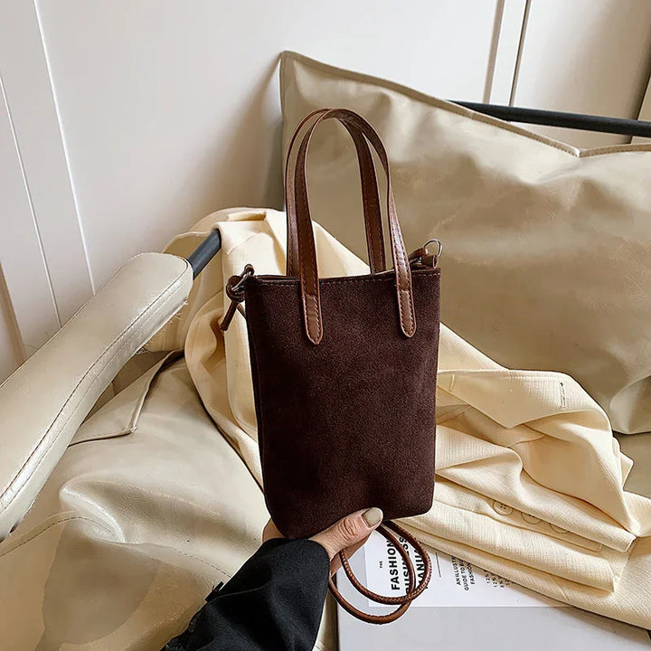 Hand holding a small dark brown suede handbag with leather handles over cream fabric and chair