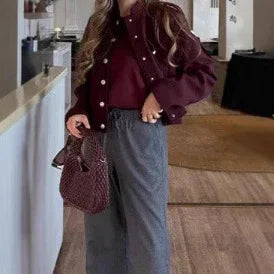 Woman wearing burgundy jacket and top with high-waisted grey pants holding a burgundy textured handbag indoors