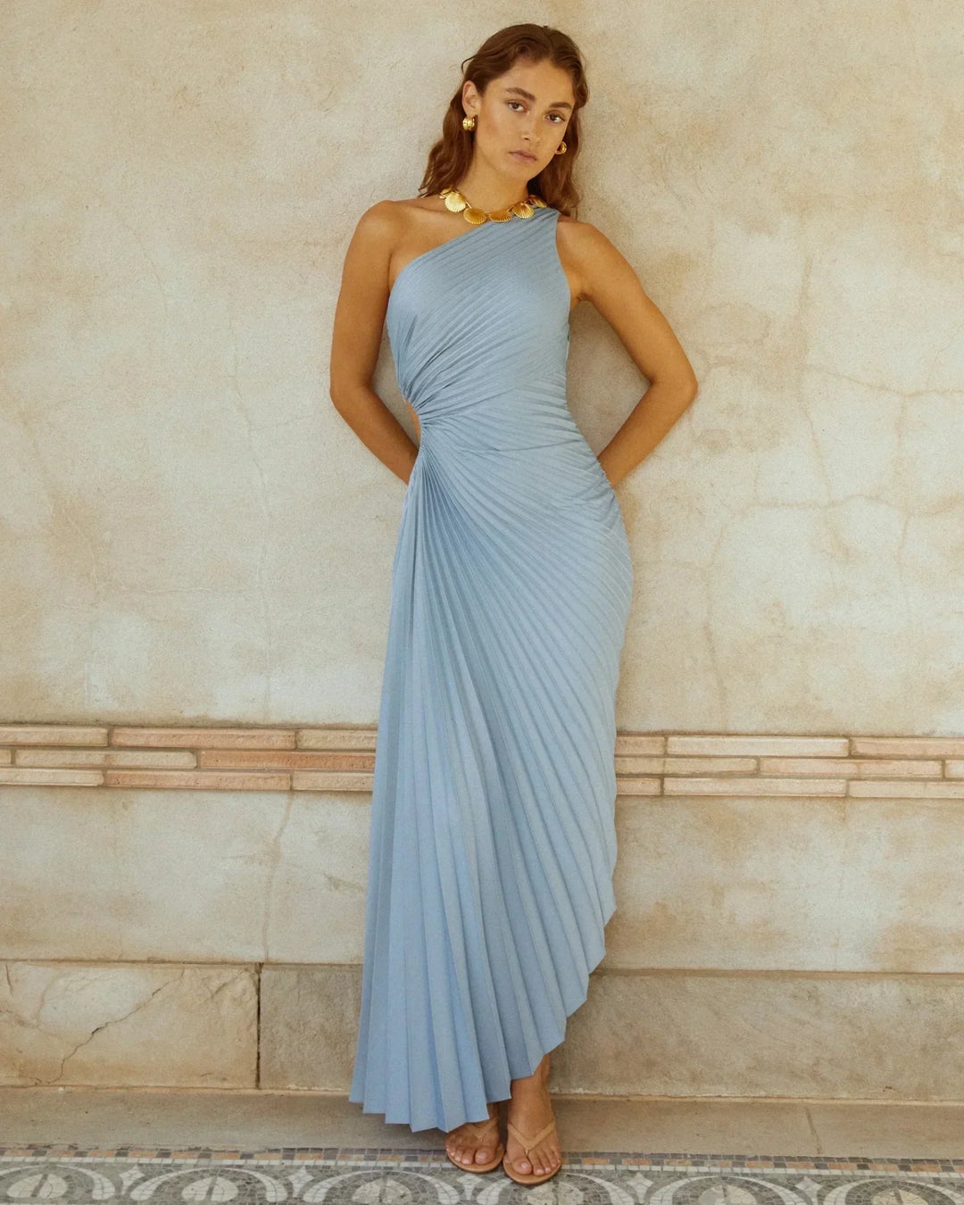 Woman in one-shoulder light blue pleated dress with gold shell necklace and earrings, standing against textured wall