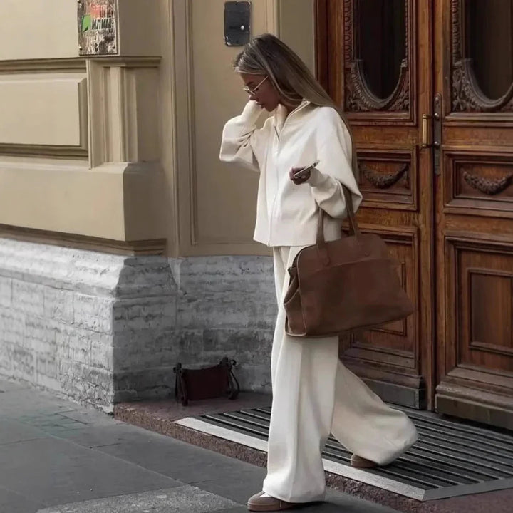 Woman in cream outfit with brown leather bag walking past ornate wooden door in city street