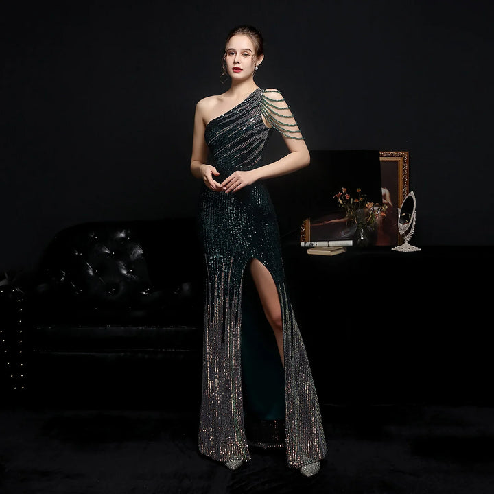 Woman in sparkly one-shoulder evening gown with beaded sleeve and thigh slit, standing by black sofa