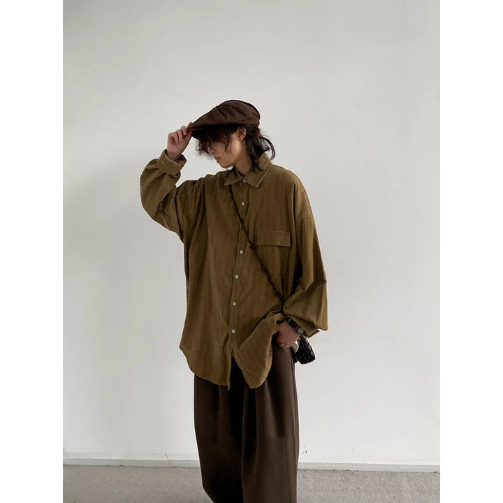 Woman wearing oversized brown shirt, wide-legged pants, and matching cap against plain wall