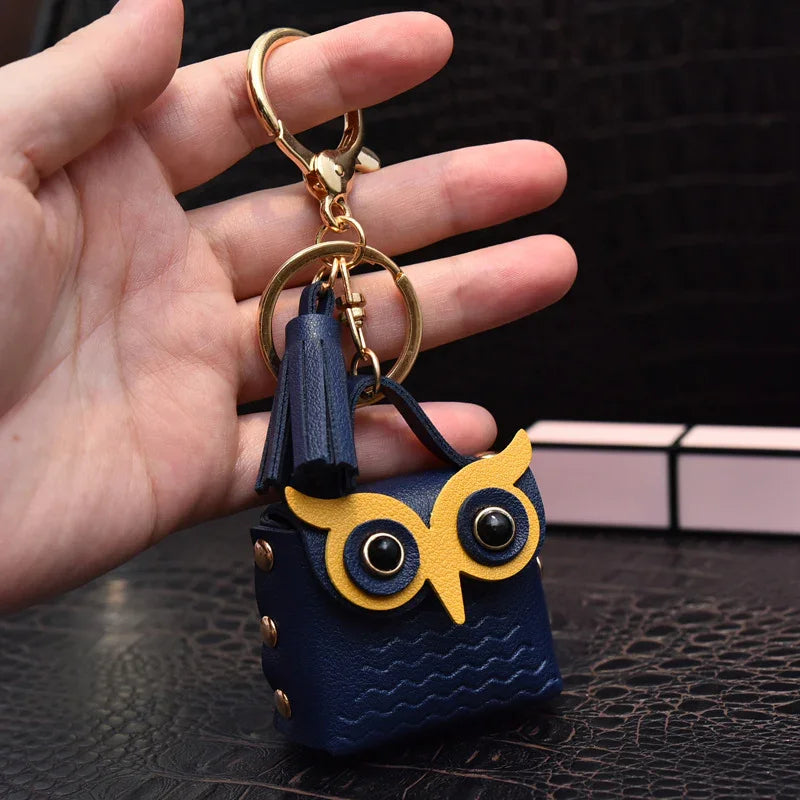 Hand holding a navy blue leather owl keychain with yellow eyes and gold hardware on black textured surface