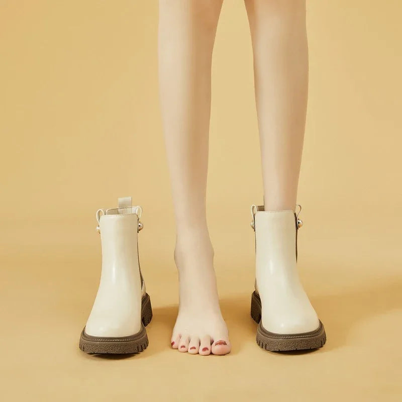 Pair of beige ankle boots with chunky brown soles and a bare foot between them on a tan background