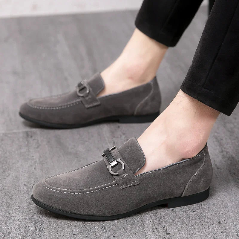 Men's gray suede loafers with metal buckle detail worn with black pants on gray floor