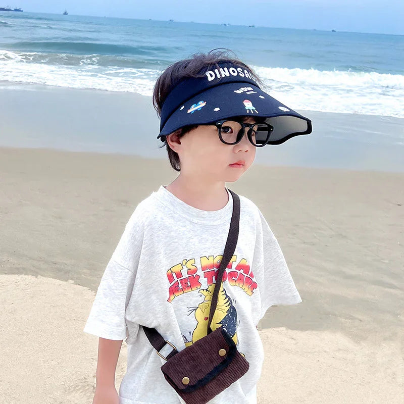 Young child wearing dinosaur visor and glasses on sandy beach with ocean waves in background