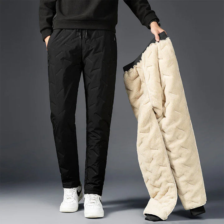 Man wearing black insulated pants and holding beige fleece-lined winter pants against gray background