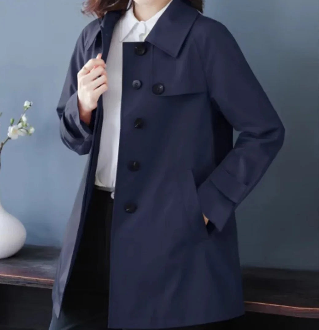 Woman wearing a navy blue buttoned trench coat over a white shirt, posed indoors with a vase and flower on a table in the background