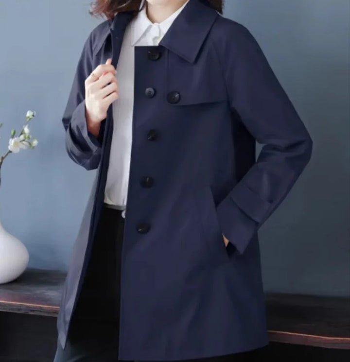 Woman wearing a navy blue buttoned trench coat over a white shirt, posed indoors with a vase and flower on a table in the background