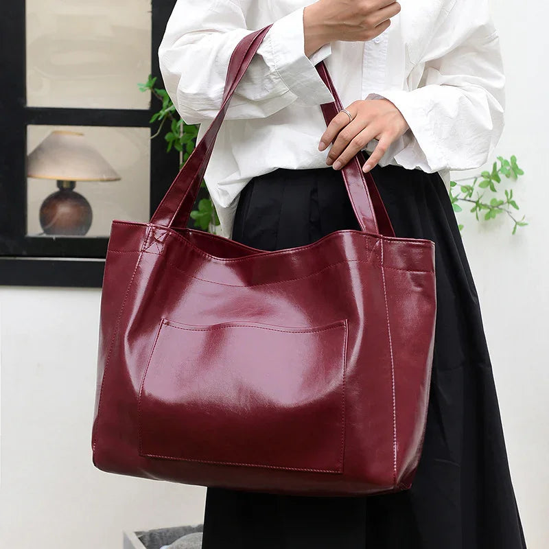 Woman holding large glossy burgundy leather tote bag with front pocket, wearing white blouse and black skirt