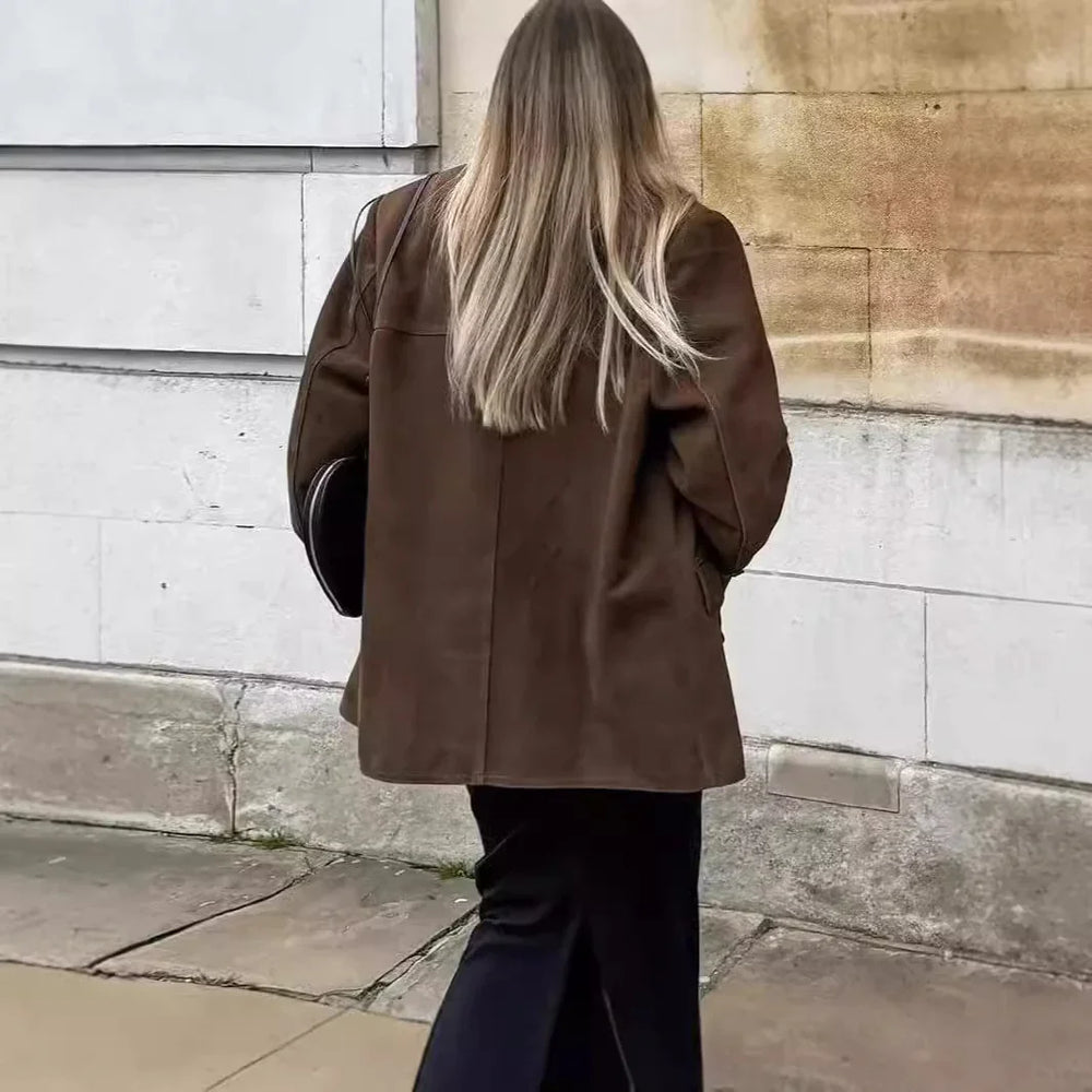 Woman with long blonde hair wearing brown jacket and black pants walking by stone wall