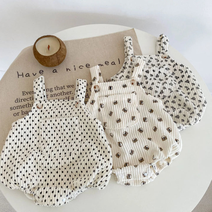 Three patterned baby rompers with straps on white table next to round candle and beige cloth