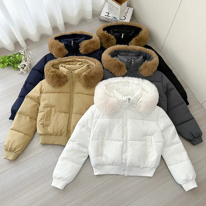 Four padded winter jackets with fur-trimmed hoods in white, beige, gray, and black colors laid out on light wooden floor