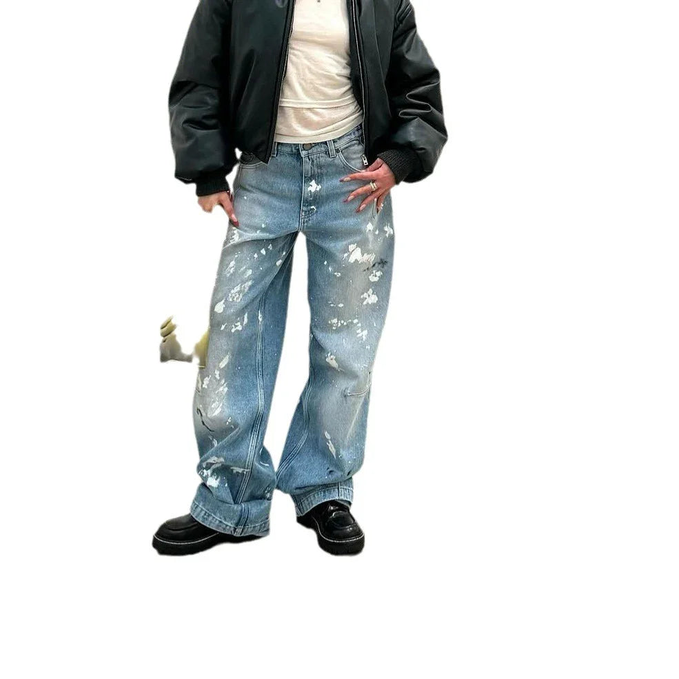 Person wearing black bomber jacket, white top, and loose-fit distressed blue jeans with paint splatters