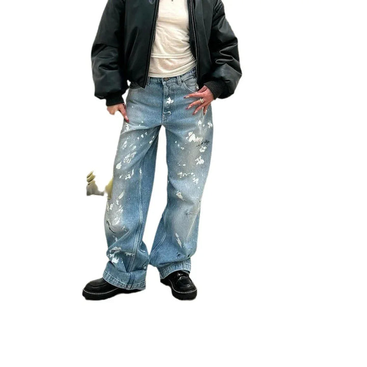 Person wearing black bomber jacket, white top, and loose-fit distressed blue jeans with paint splatters
