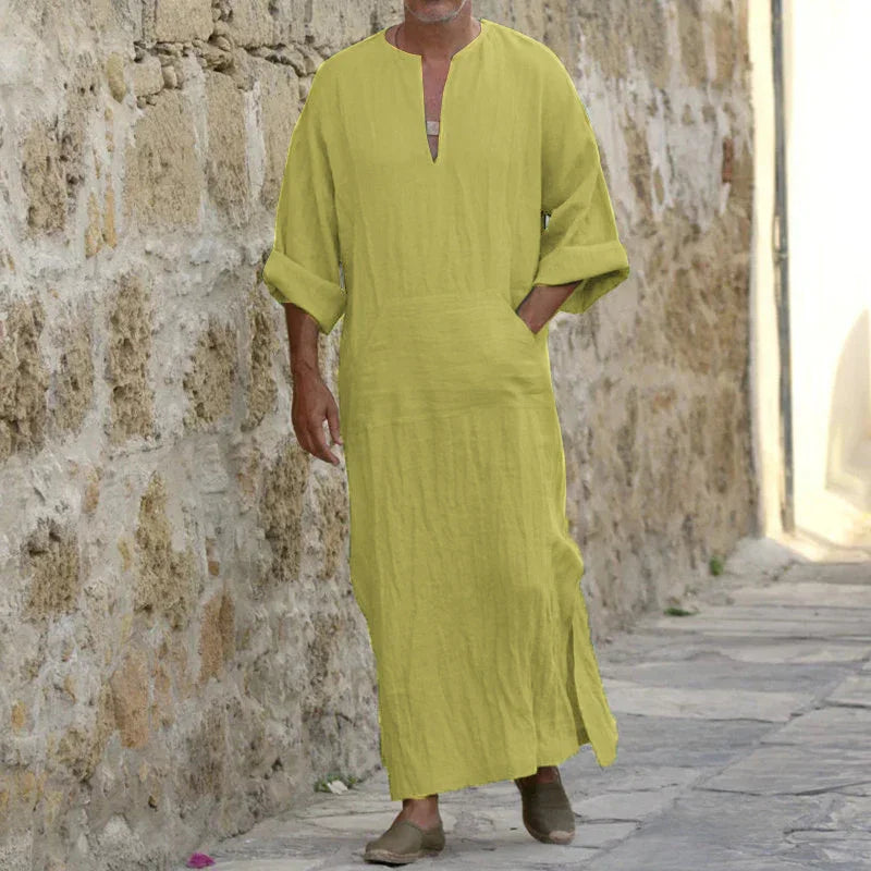 Man wearing a loose, yellow linen tunic dress walking beside a stone wall on a cobblestone street