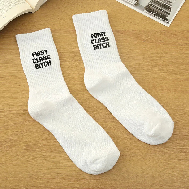 Pair of white ribbed socks with black text 'FIRST CLASS BITCH' on wooden surface, surrounded by books