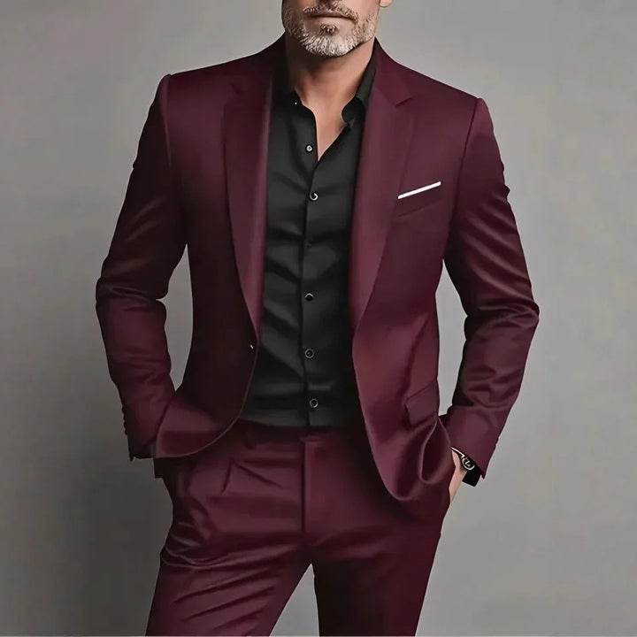 Man wearing a tailored burgundy suit with black dress shirt and white pocket square against gray background