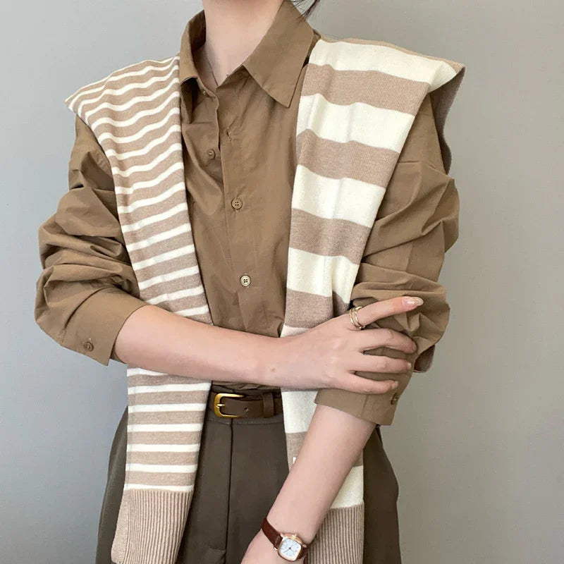 Woman wearing brown shirt with rolled-up sleeves, striped beige and white sweater draped over shoulders, brown belt, and wristwatch