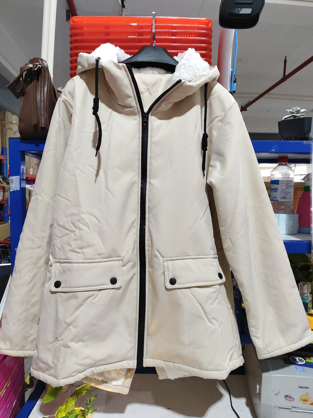 Beige hooded winter coat with black zipper and large front pockets, displayed indoors