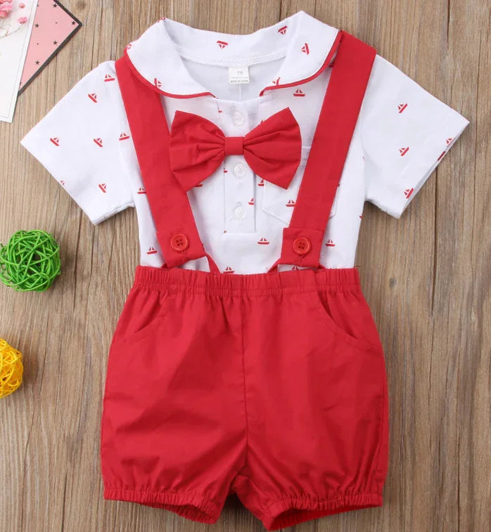 Baby boy outfit with white shirt featuring red sailboat prints, red bow tie, and red suspenders with shorts on wooden background