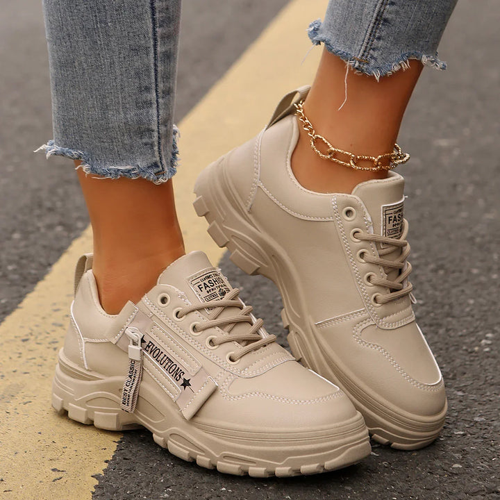 Beige chunky sneakers with EVOLUTIONS tag worn with frayed hem jeans and gold chain anklet on asphalt