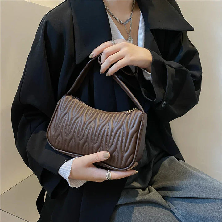Woman holding brown quilted leather handbag with wavy stitching, wearing black coat and silver jewelry