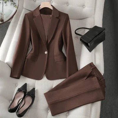 Brown tailored women's business suit with blazer and pants, paired with black pointed-toe heels and a black handbag on tufted beige chair
