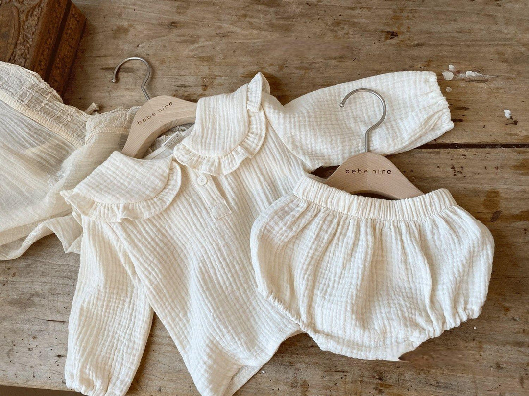 Soft cream-colored baby outfit with ruffled collar and matching bloomers on wooden hangers