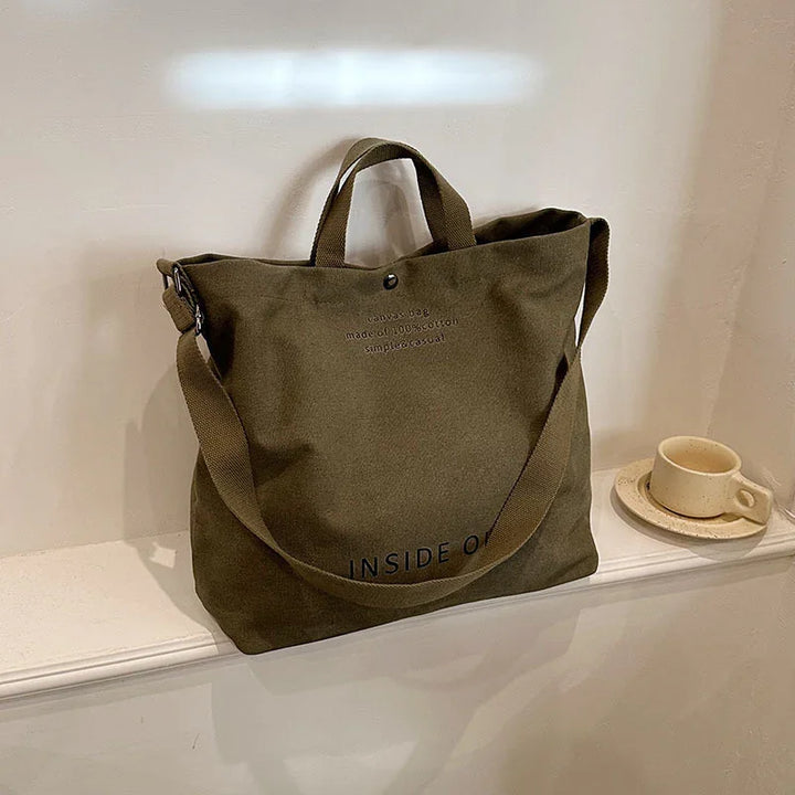 olive green canvas tote bag with handles and shoulder strap on white ledge next to ceramic cup and saucer