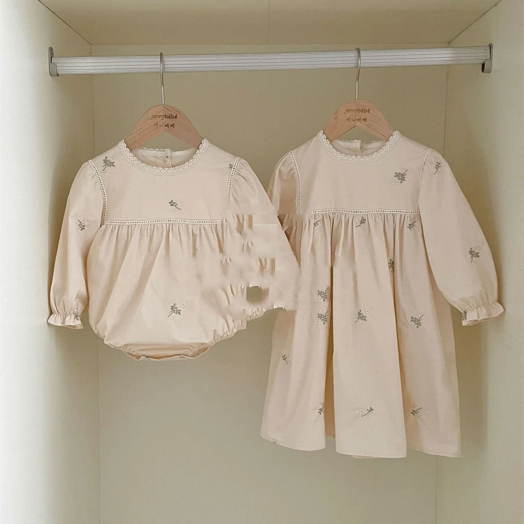 Beige baby romper and dress with floral embroidery hanging on wooden hangers in closet
