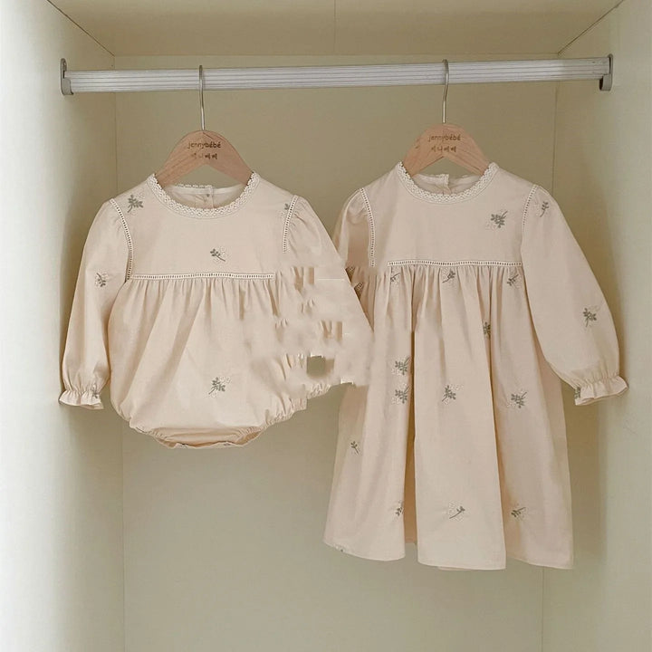 Beige baby romper and dress with floral embroidery hanging on wooden hangers in closet