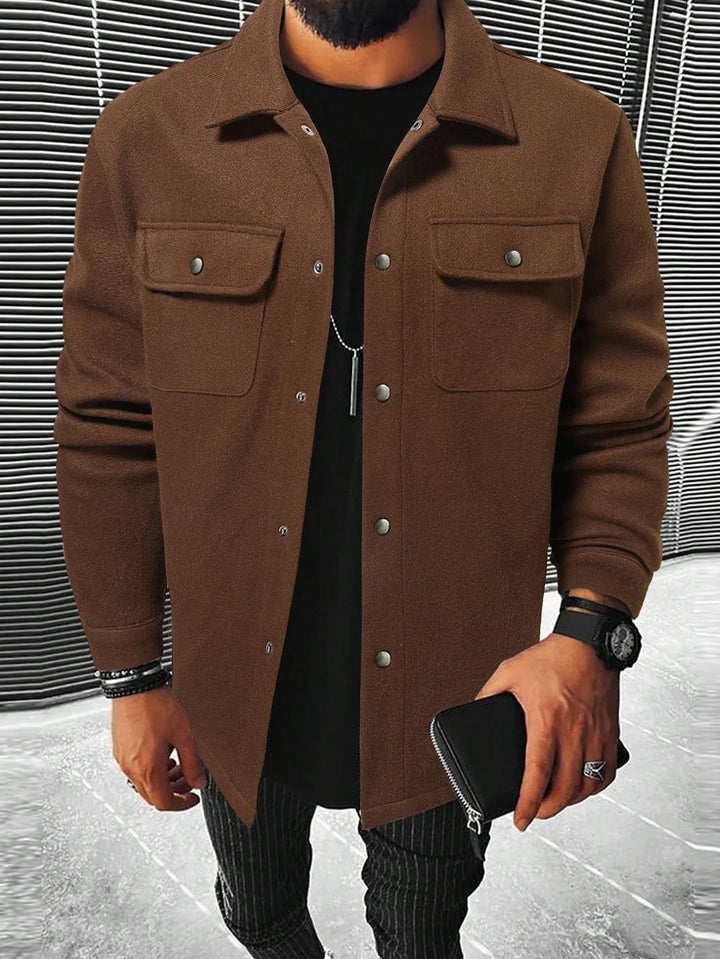 Man wearing brown button-up jacket with flap pockets, black shirt, striped pants, and holding black wallet