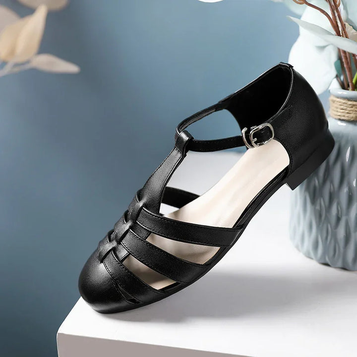 Black leather women's flat sandal with T-strap and buckle on white surface next to blue vase