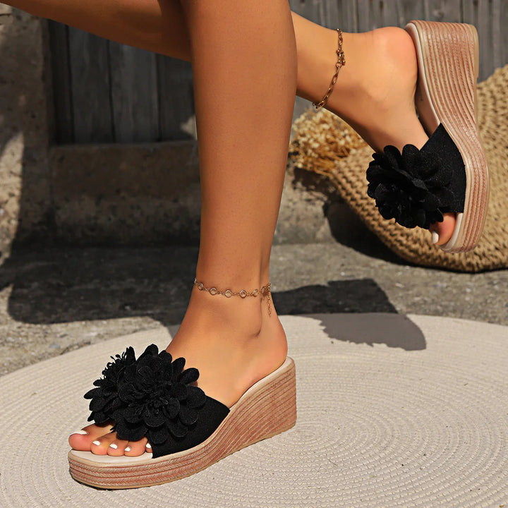 Black floral wedge sandals on feet with gold anklets in outdoor setting with woven mat