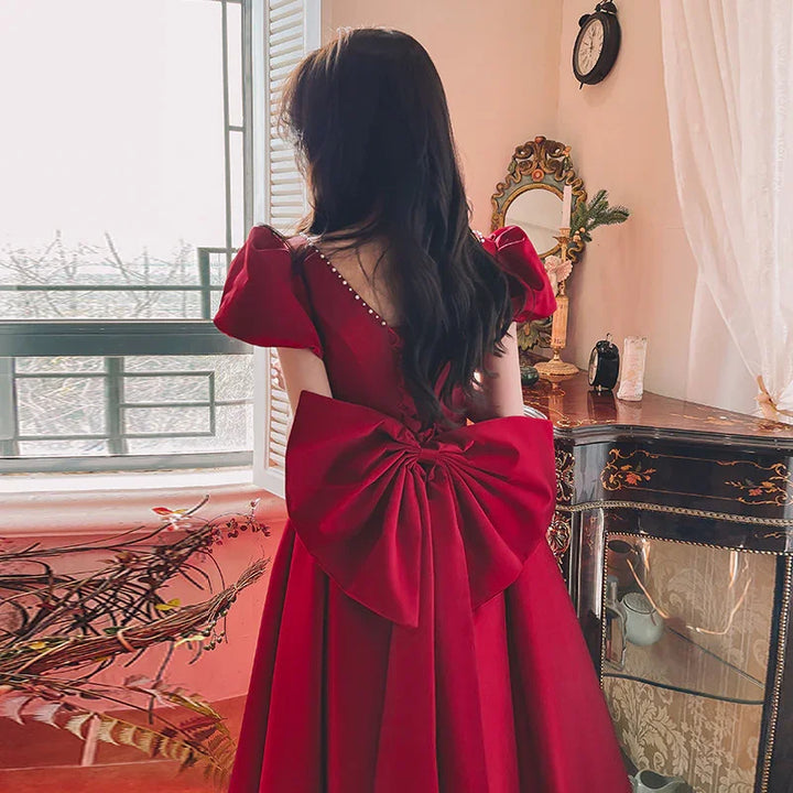 Woman in elegant red dress with large bow back detail standing by vintage black dresser near window