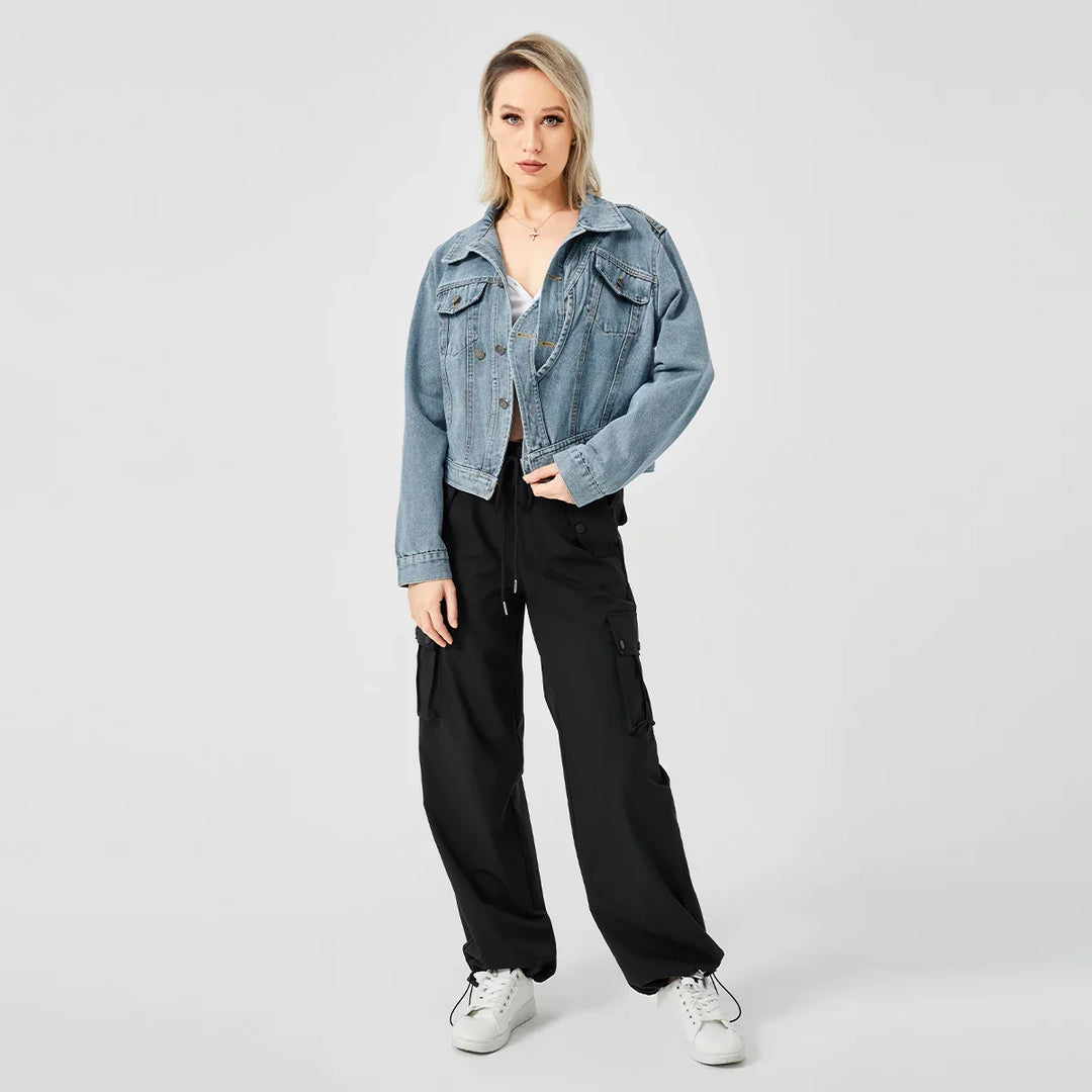 Blonde woman wearing blue denim jacket, black cargo pants, and white sneakers on gray background