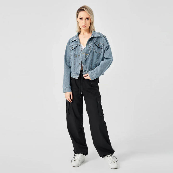 Blonde woman wearing blue denim jacket, black cargo pants, and white sneakers on gray background