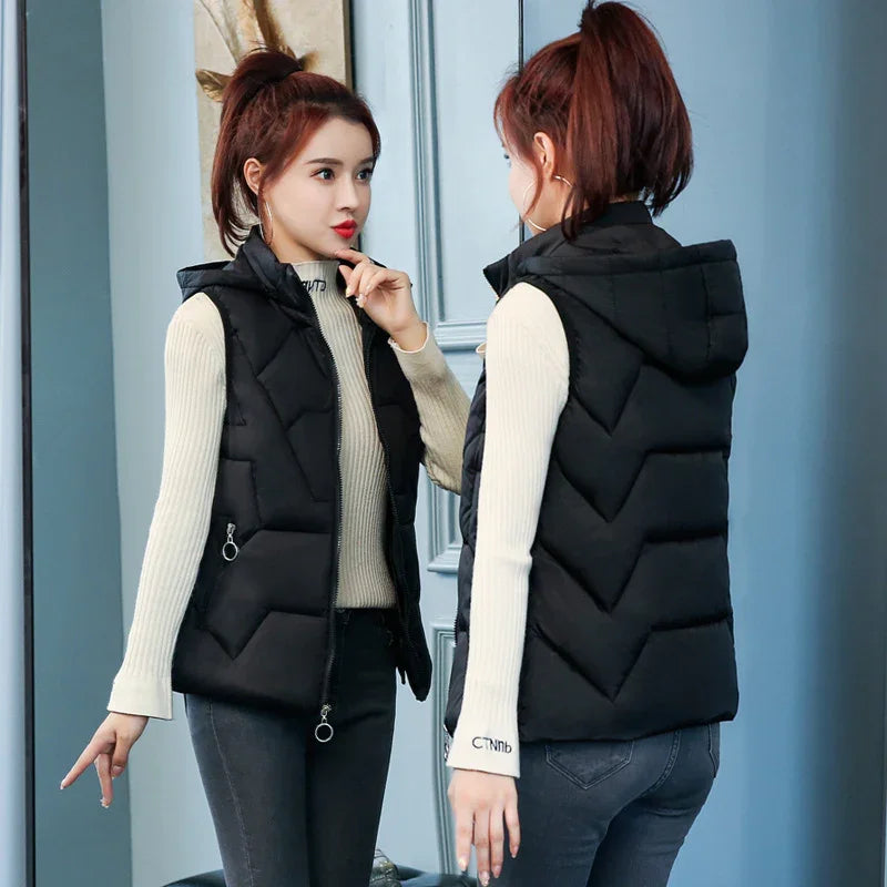 Woman wearing black hooded quilted vest and beige ribbed sweater, standing by a mirror